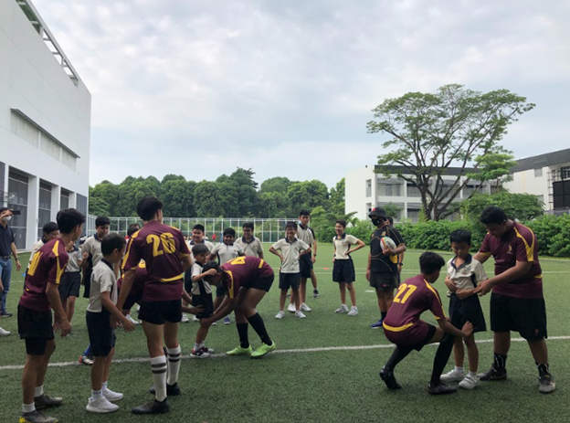 Students in maroon and yellow jerseys practice rugby drills on a green field beside school buildings and trees.